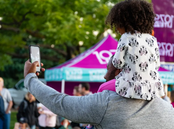 father holds daughter on shoulders at an event and takes a selfie